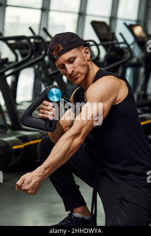 Ein junger Mann benutzt und genießt die Schlagmassagepistole, um sich zu entspannen und die Schmerzen im Körper und in den Muskeln im Fitnessstudio zu lindern. Stockfoto