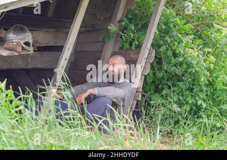 Viking Krieger ruhen in der Nähe von Holzgebäude nach der Schlacht. Metallkettenpost und Helm. Grüne Büsche. Stockfoto