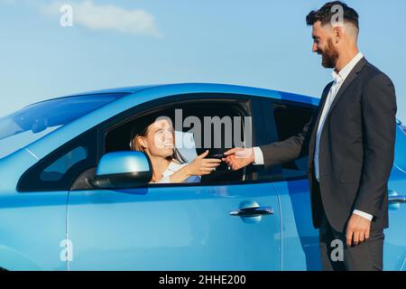 Der männliche Autoverkäufer übergibt die Schlüssel an die Käuferin, der Kaufvertrag ist erfolgreich abgeschlossen Stockfoto