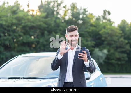 Glücklicher Geschäftsmann Autoverkäufer inspiziert das Auto online mit einem Videoanruf und einer Anwendung am Telefon, ein Mann hat Spaß, ein Auto zu verkaufen Stockfoto