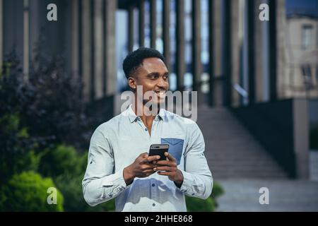 Junge glückliche afroamerikanische männliche Freiberufler korrespondieren in Handy und lesen gute Nachrichten aus Handy online Stockfoto