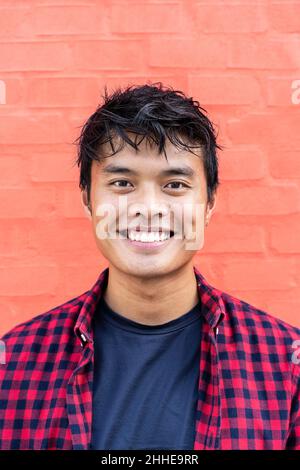 Porträt eines hübschen jungen asiatischen Mannes, der im Freien vor einem roten Wandhintergrund in der Stadt steht und die Kamera anschaut - Happy Asian guy Portrait - Gattungen Stockfoto
