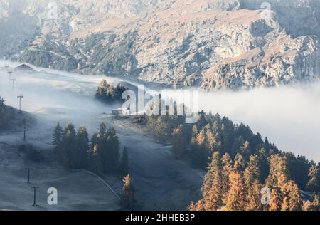 Morgennebel zieht über gefrorene Hänge, goldene Lärchen und Skilifte unter felsigen Dolomitenklippen auf der Alpe di Siusi, Südtirol, Italien. Stockfoto