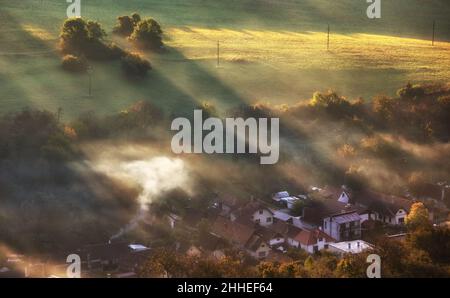 Luftaufnahme des Dorfes im Nebel mit goldenen Sonnenstrahlen bei Sonnenaufgang im Herbst. Schöne ländliche Landschaft mit Straße, Gebäuden, nebligen bunten Bäumen. Slovaki Stockfoto