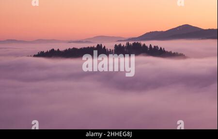 Sonnenuntergang versteckt hinter Wolken und Nebel über Hügeln, leuchtend gelbe Sonne auf bunte Wolkenlandschaft, blau violett orange Himmel. Stockfoto