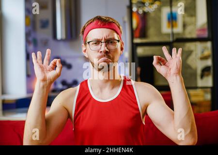 Porträt eines lustigen Sportlers in roter Sportkleidung, der das OK-Zeichen auf dem Sofa zu Hause macht. Stockfoto