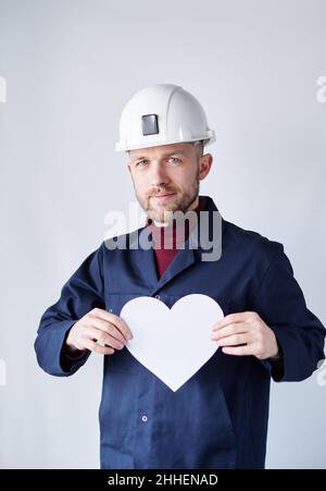 Romantik, Liebe oder st. Valentinstag-Konzept. Porträt eines jungen glücklichen bärtigen männlichen Arbeiters im Helm, der an der Wand steht und die Herzform aus weißem Papier lächelt. Hochwertiges vertikales Bild Stockfoto