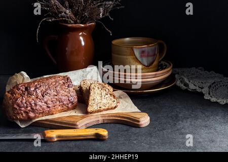 Brot mit Sonnenblumen- und Leinsamen auf einem Holzschneidebrett. Keramikgeschirr im Hintergrund. Stockfoto
