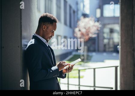 Junger afroamerikanischer Geschäftsmann im formellen Business-Anzug stehend mit Tablet in den Händen auf dem Hintergrund modernes Bürogebäude draußen arbeiten. Mann usi Stockfoto