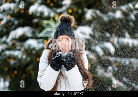 Die junge Frau bläst Schnee aus ihren Händen, während sie vor dem Hintergrund eines Weihnachtsbaums steht Stockfoto