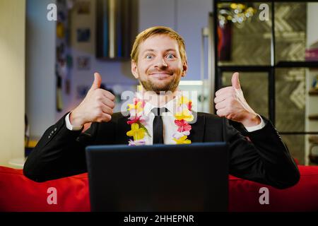 Junger Geschäftsmann in Anzug und hawaiianische Halskette sitzt in seinem Home Office mit Daumen nach oben und mit Laptop. Urlaub in einer Pandemie. Stockfoto