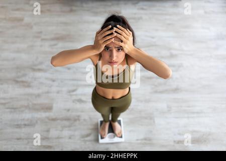 Erschrocken junge indische Frau, die auf einer Waage steht, den Kopf in Enttäuschung haltend, ohne ein Gewichtsverlust Ergebnis zu erzielen Stockfoto