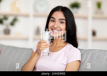 Lächelnde junge Inderin, die ein Glas frisches, sauberes Wasser in der Hand hält, während sie auf der Couch im Wohnzimmer sitzt. Halten Sie hydratisiert Konzept Stockfoto