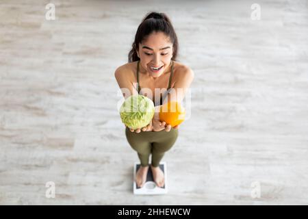 Oben sehen Sie die tausendjährige indische Dame, die Zitrusfrüchte und Kohl hält und sich für eine gesunde Ernährung im Haus entscheidet Stockfoto