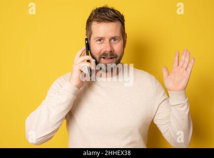 Junger Mann im lässigen Pullover, der auf dem Smartphone spricht und mit einem überraschten Gesicht auf gelbem Hintergrund auf die Kamera schaut. Stockfoto