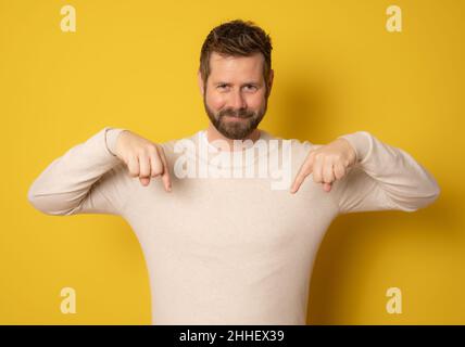 Junger Mann isoliert auf gelbem Hintergrund zeigt mit Fingern nach unten, positives Gefühl. Stockfoto