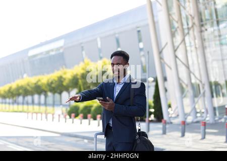 afroamerikanischer Geschäftsmann, der auf dem Hintergrund eines modernen Bahnhofsflughafens in formellem Anzug mit einem Koffer per App-Handy steht. Traveli Stockfoto