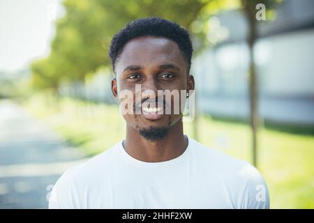 Porträt eines jungen, tausendjährigen afroamerikanischen Mannes, der glücklich und lächelnd in einem weißen T-Shirt auf die Kamera blickt Stockfoto