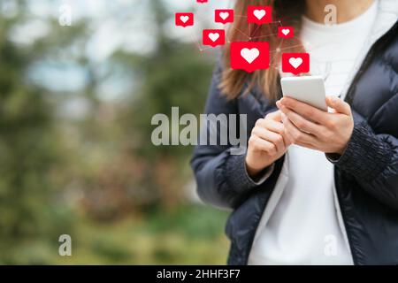 Interaktionen in sozialen Medien auf dem Mobiltelefon. Junge Frau hält das Smartphone mit „Gefällt mir“- und „Liebe“-Symbolen, während sie im Freien steht Stockfoto