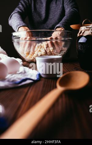 Frau Hände Kneten Teig für Kuchen in großen Glasschüssel auf braunem Holztisch, Nahaufnahme Stockfoto