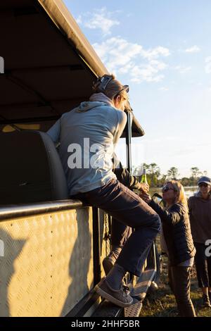 Eine Frau, die mit einer Flasche Wein am Rand eines Jeeps sitzt und einen Sundowner hat. Stockfoto
