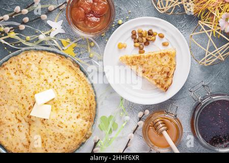 Stapel von hausgemachten frittierten Crepes mit Butter, Gläsern mit Honig, Himbeermarmelade, Aprikosenmarmelade und Weihnachtsdekoration auf blauem Strukturhintergrund. Russisch Stockfoto