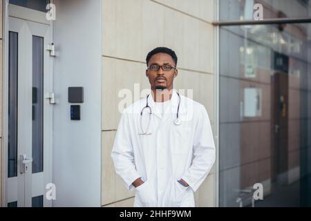 Porträt eines jungen, ernsthaften afro-amerikanischen Arztes in der Nähe der Klinik in einem weißen medizinischen Gewand, der auf die Kamera schaut Stockfoto