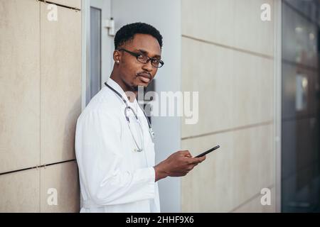 Ein seriöser afroamerikanischer Arzt in der Nähe der Klinik, in einem weißen medizinischen Gewand, blickt auf die Kamera und hält ein Mobiltelefon, um mit den Patienten zu sprechen Stockfoto