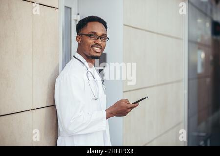 Ein fröhlicher Mann eines afroamerikanischen Arztes, in der Nähe der Klinik, in einem weißen medizinischen Gewand, blickt auf die Kamera und hält ein Mobiltelefon, um mit dem Patienten zu sprechen Stockfoto