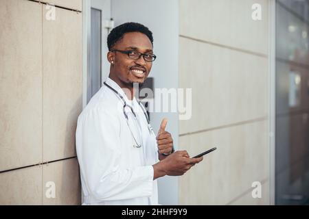 Ein fröhlicher Mann eines afroamerikanischen Arztes, in der Nähe der Klinik, in einem weißen medizinischen Gewand, blickt auf die Kamera und hält ein Mobiltelefon, um mit dem Patienten zu sprechen Stockfoto