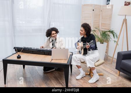 Lächelnde afroamerikanische Frau spielt akustische Gitarre neben Freund mit Synthesizer und Laptop im Wohnzimmer Stockfoto