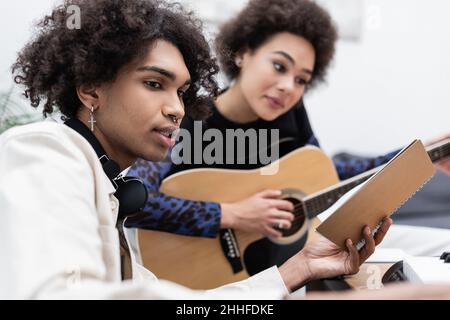 afroamerikanischer Mann mit Kopfhörern, der das Notebook in der Nähe einer verschwommenen Freundin hält, die zu Hause akustische Gitarre und Synthesizer spielt Stockfoto