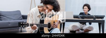 Junger afroamerikanischer Mann spielt akustische Gitarre in der Nähe verschwommene Freundin im Wohnzimmer, Banner Stockfoto