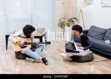 Lächelndes afroamerikanisches Paar, das im Wohnzimmer akustische Gitarre und Synthesizer spielt Stockfoto