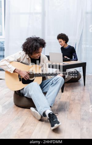 Lächelnde afroamerikanische Frau spielt Synthesizer in der Nähe von Laptop und Freund mit Akustikgitarre im Wohnzimmer Stockfoto