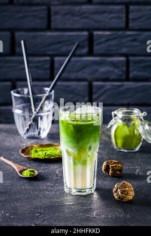 Iced Green Matcha Tee oder Matcha Latte gemischt mit Eiswürfel und Milch in einem hohen Glas mit einer Ziegelwand im Hintergrund mit gepudertem Matcha in Glas j Stockfoto