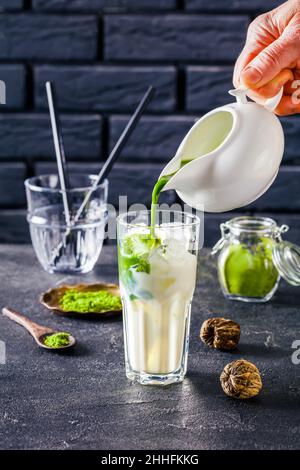 Barista gießt Matcha-Tee von Hand in ein Glas mit Milch gemischt mit Eiswürfeln mit einer Backsteinwand im Hintergrund und Matcha-Tee in Pulverform in einem Glas, cl Stockfoto