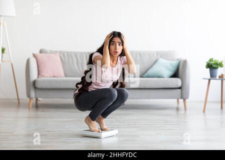 Schockierte junge indische Dame, die auf einer Waage sitzt, erschreckt vor Gewichtszunahme zu Hause, freiem Platz. Gesundes Wohnkonzept Stockfoto