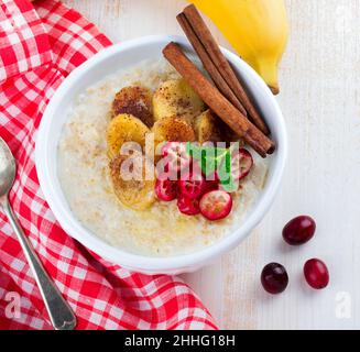 Haferbrei mit gebackenen Bananen, Zimt, Orange und Preiselbeeren in einer Keramikschale für ein gesundes Frühstück auf hellem Hintergrund.Draufsicht. Selektiver FOC Stockfoto