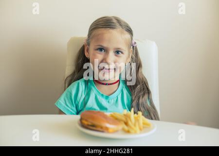 Ein lächelndes kleines Mädchen sitzt am Tisch mit Hamburgern und Pommes Frites Stockfoto