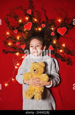 Babymädchen mit roten Herzen in lockigem Haar liegt und lächelt mit einem Teddybären in den Händen. Valentinstag Stockfoto