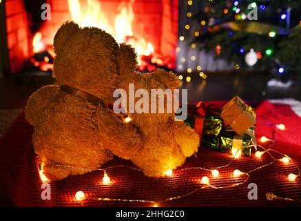 Zwei niedliche Teddybären umarmen sich gegenseitig, sitzen auf einem gestrickten roten Karree vor dem Hintergrund eines Weihnachtsbaums, von Geschenken und einem brennenden Kamin. Stockfoto