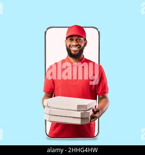 Fröhlicher junger schwarzer Lieferer in Uniform gibt viele Kartons Pizza vom leeren Bildschirm des großen Telefons Stockfoto
