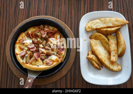 Barbeque-Pizza-Hütte mit Rind, serviert auf einem schwarzen Teller oder einer Schüssel mit einem braunen strukturierten Holztisch im Hintergrund. Gewürzte Bratkartoffeln auf einem weißen Teller serviert. Stockfoto