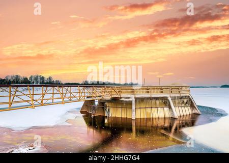 Gefrorener Fluss mit Staudamm am wunderschönen Sonnenaufgang. Blick auf den im Winter mit Eis bedeckten Staudamm. Stockfoto