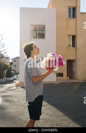 Ein junger Mann, der in einen Blumenstrauß aus Bougainvillea verliebt ist, steht in der Nähe des Gebäudes und schaut nach oben. Valentinstag. Eine angenehme Überraschung für ein Mädchen, Mutter, Stockfoto