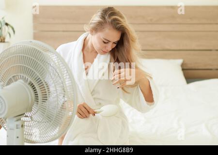 Junge Frau mit langen blonden Haaren sitzt vor einem Ventilator und bürstet Haare Stockfoto