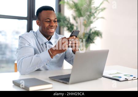 Gutaussehender, selbstbewusster junger afroamerikanischer Geschäftsmann, Agent, Manager, sitzt am Arbeitsplatz im modernen Büro, nutzt Smartphone, chattet am Telefon mit Kollegen oder Kunden, lächelt freundlich Stockfoto