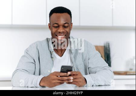 Afroamerikanischer attraktiver positiver junger Mann, in stilvoller Freizeitkleidung, verbringen Freizeit am Tisch in der Küche sitzend, nutzt Smartphone, stöbert im Internet, chattet in sozialen Medien, lächelt Stockfoto