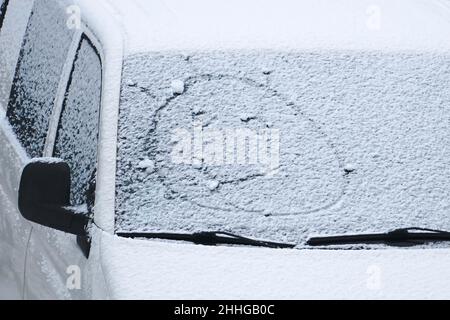 Ein gezeichnetes Smiley-Gesicht im Winter auf der schneebedeckten weißen Autoscheibe. Eine grafische Kreativszene für die Wintersaison. Der positive Ausdruck als lustig Stockfoto